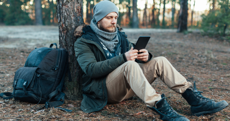 A man taking a break from hiking, sitting against a tree and reading a kindle