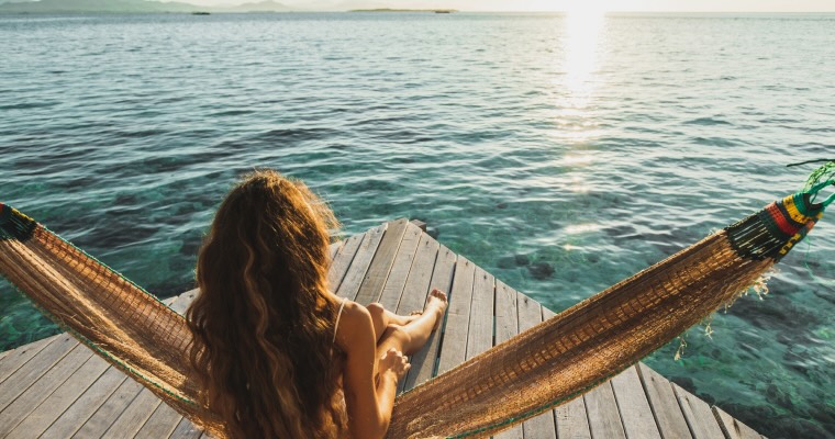 View from behind of woman awakening with ocean view. Wooden hotel terrace with hammock. Beautiful morning light