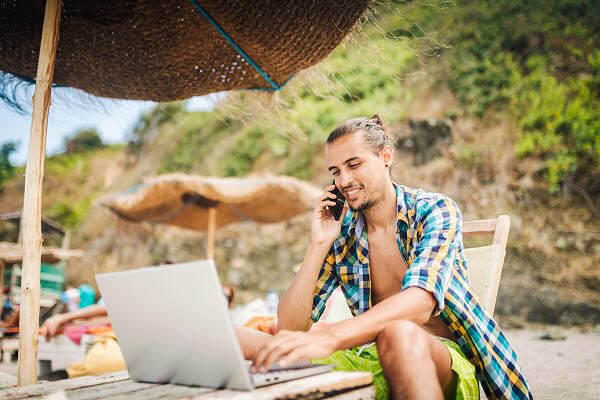 A man on the phone and working on a laptop while sitting on a lounger chair on the beach